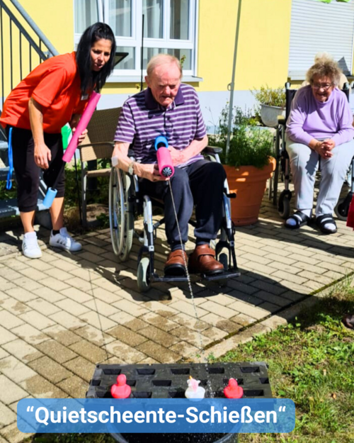 Senioren und Senioren sitzen im Rollstuhl draußen im Innenhof, begleitet von einer Betreuungskraft. Sie haben Spaß mit sommerlichen Wasserspielen. Bildbeschriftung: “Quietscheente-Schießen“